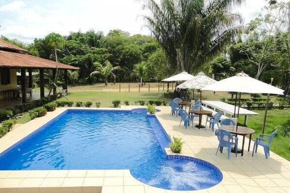 Una piscina con idromassaggio, circondata da mobili da giardino e ombrelloni, immersa in un rigoglioso giardino con una casa sullo sfondo.