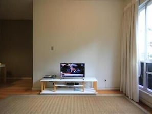 Quarto com televisão sobre um suporte branco, janela com cortinas e chão de madeira.