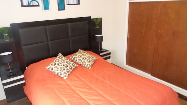 A bed with an orange bedspread and two patterned pillows, a black headboard, bedside tables with lamps, and a wooden wardrobe. - Departamento MDQ Entre Rios by EF (Mar del Plata)