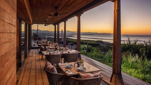 Een terras met houten vloer en tafels gedekt voor een maaltijd, met uitzicht op een schilderachtig zee- en berglandschap.