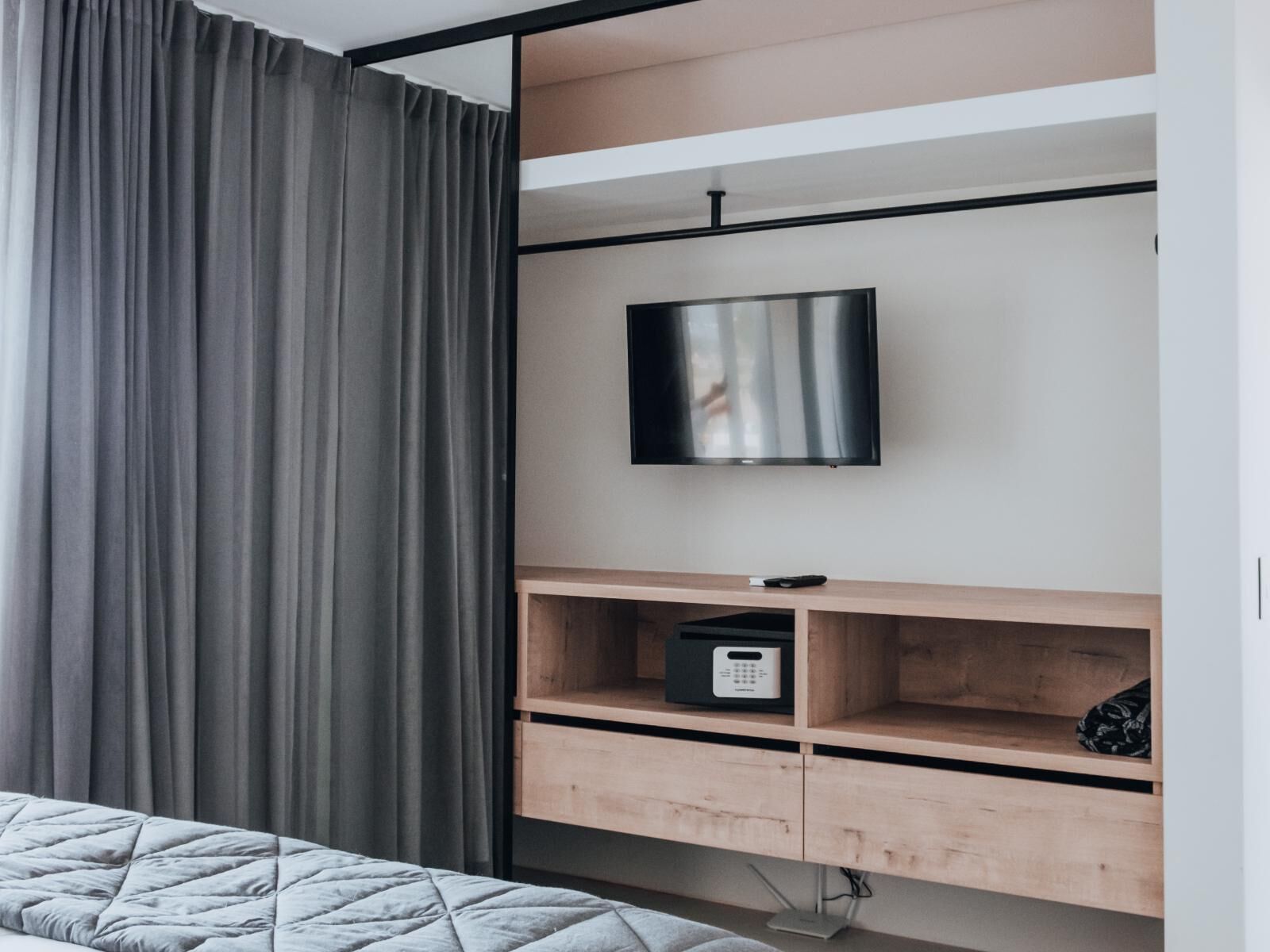 A modern bedroom with a bed, a TV mounted on a wooden shelf, and a bedside table with a remote control.