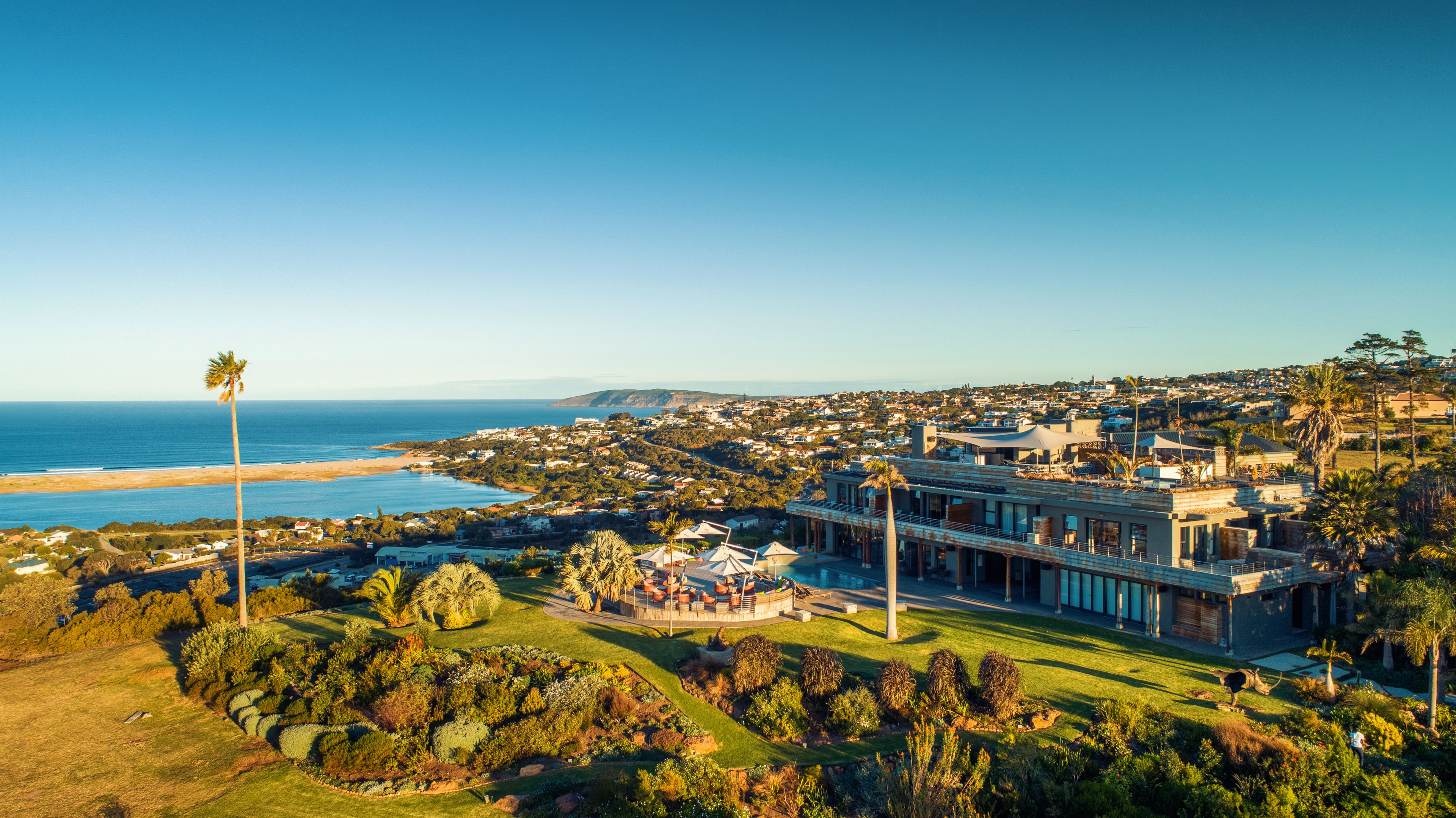 Un complejo turístico frente al mar, con piscina, jardines exuberantes y vistas al océano y colinas.