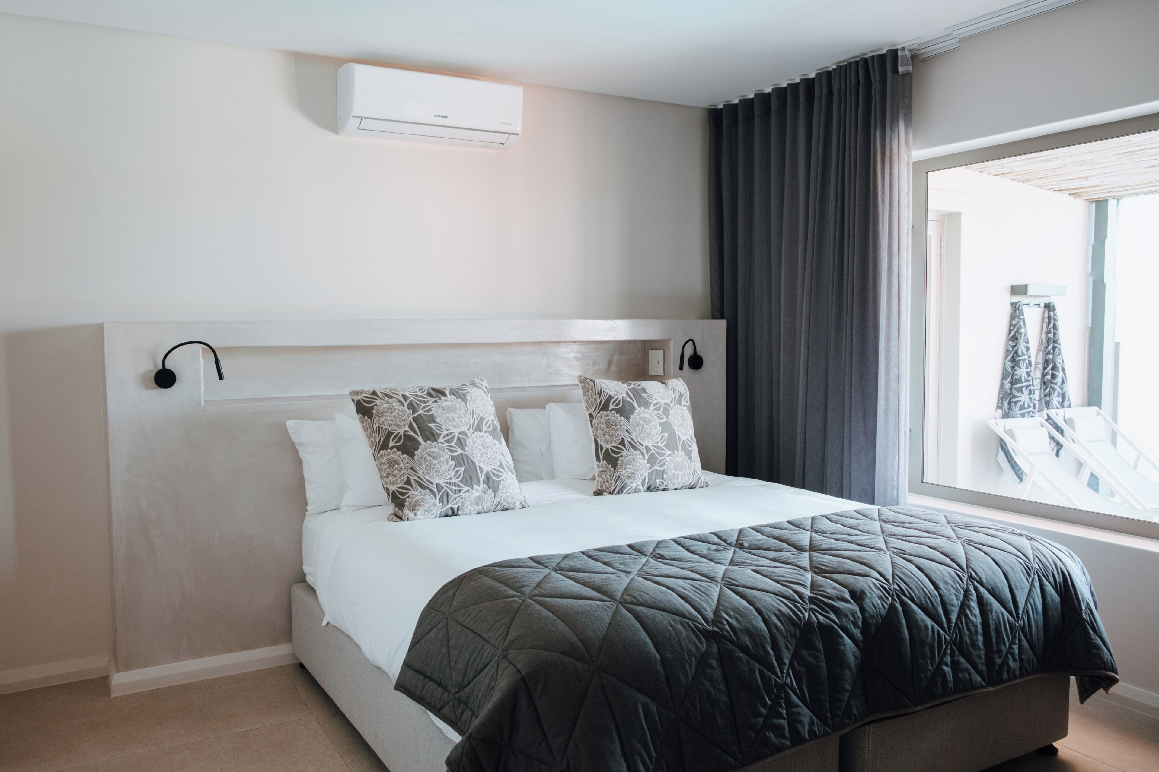 A modern hotel room with a large bed, a headboard with built-in speakers, a window with curtains, and a wall-mounted air conditioner.