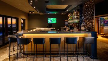 Zona de bar con mostrador, taburetes, una pantalla de televisión y una pared llena de botellas de licores.