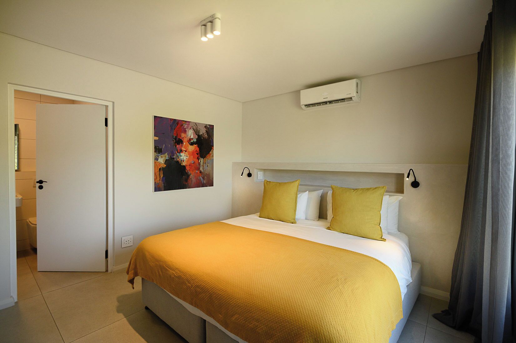 A bedroom with a bed, a painting, an air conditioning unit, and a door to the bathroom.