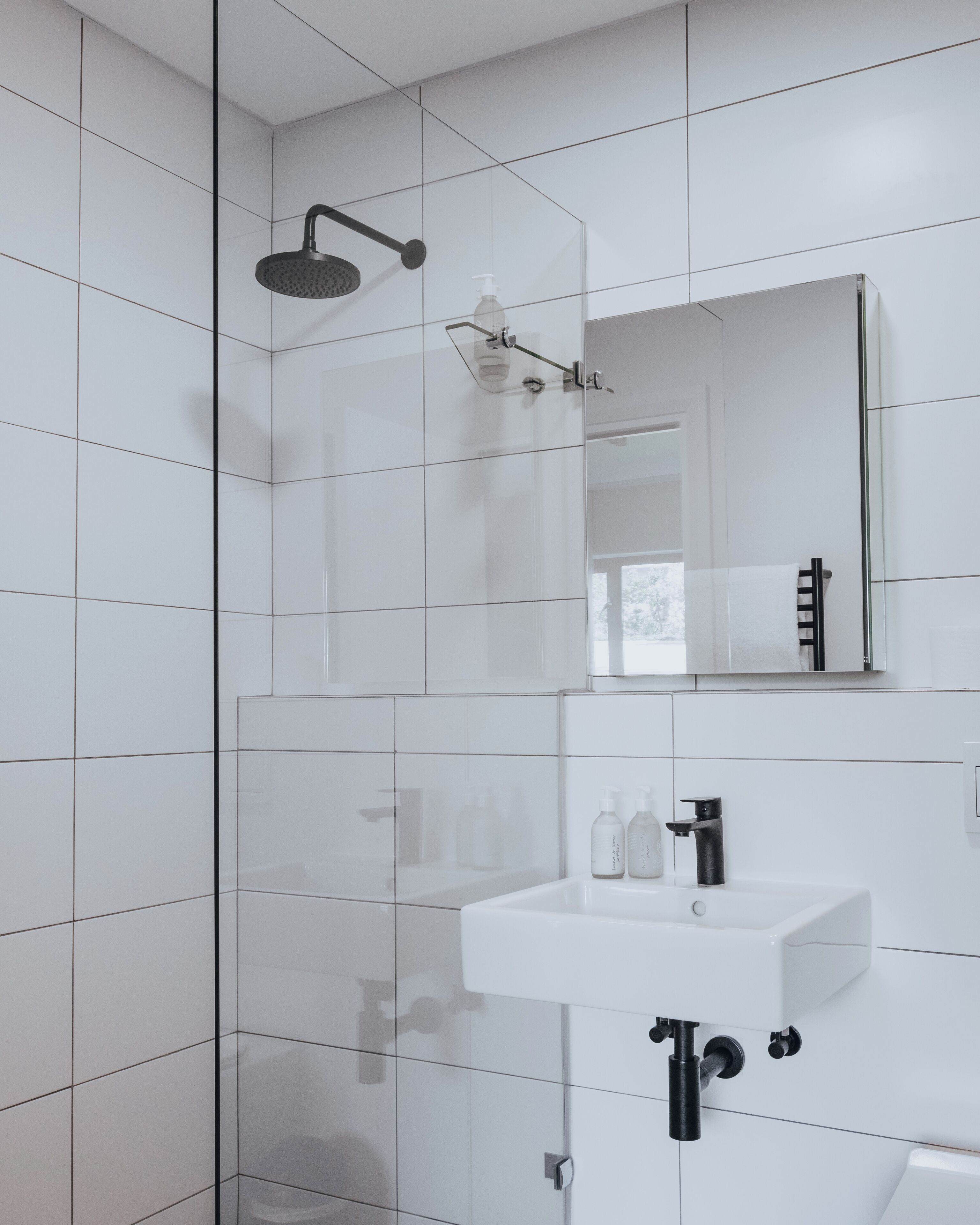 Une salle de bain moderne avec des carreaux blancs, une douche en verre, un lavabo blanc avec un robinet noir et un miroir au-dessus du lavabo.