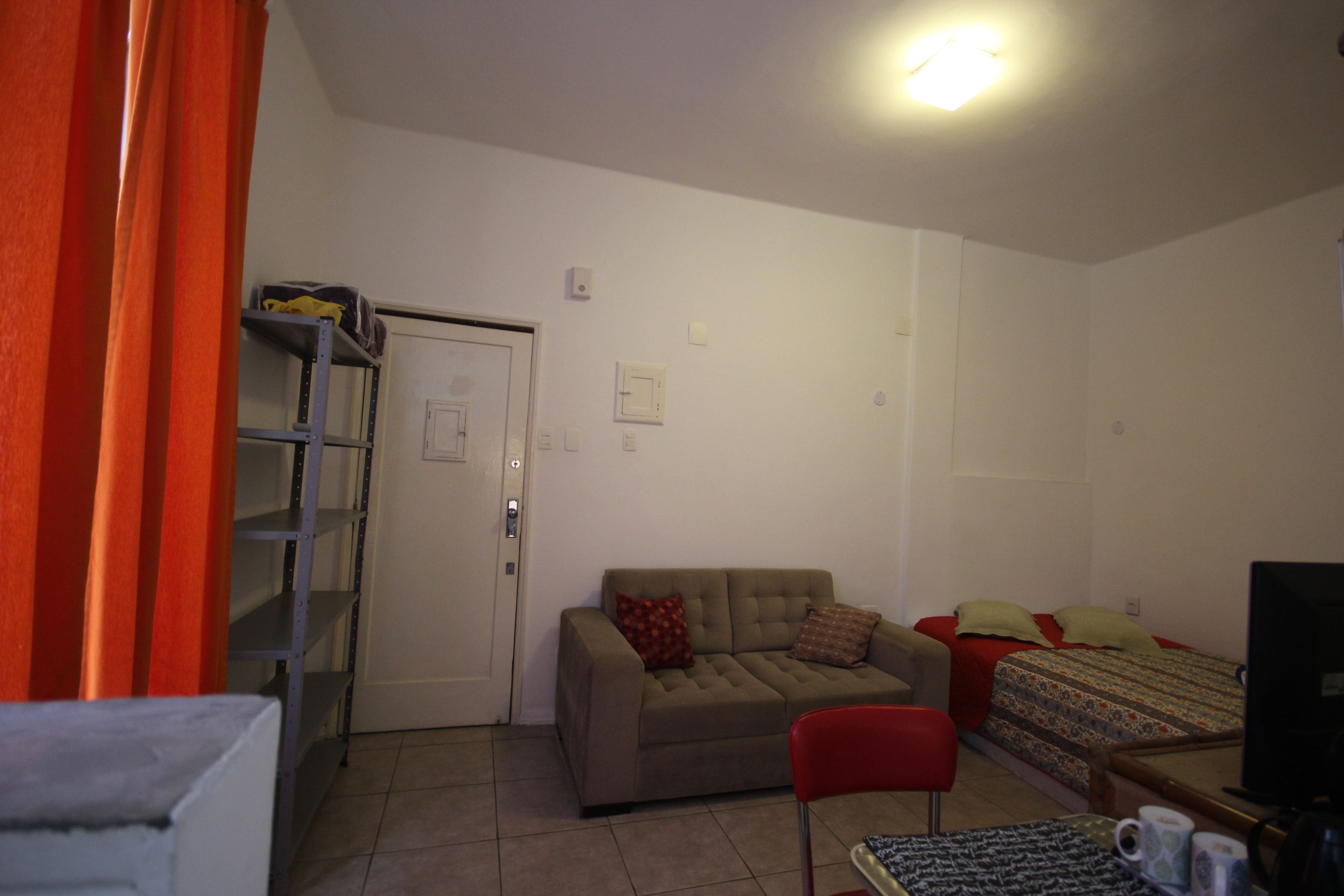 Une petite pièce bien éclairée, avec un canapé, un lit, un téléviseur et une étagère.