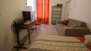 Una habitación pequeña y amoblada con una cama, un escritorio con una computadora, una silla, un sofá y un ventilador.