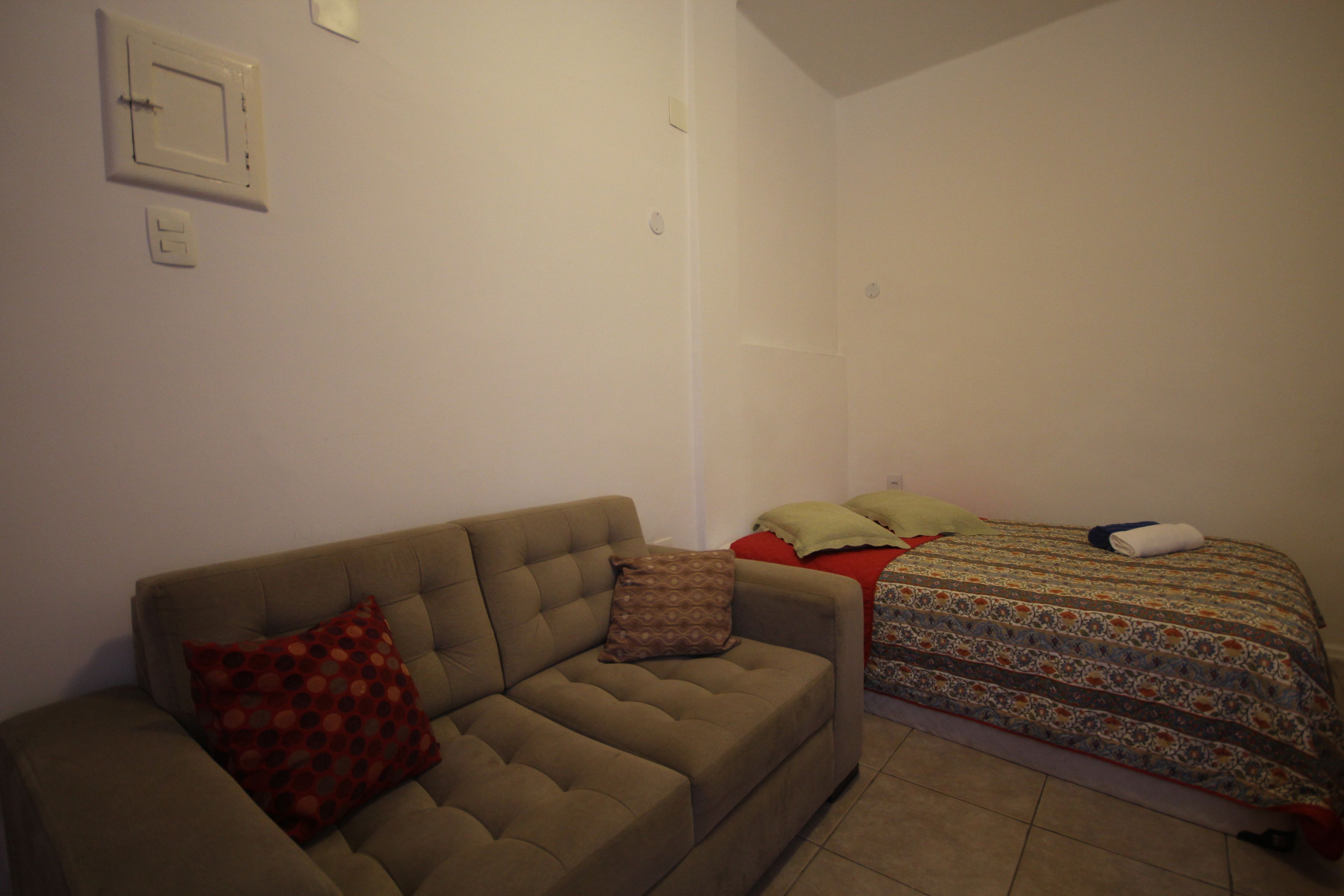 A room with a sofa, a bed with patterned bedding, and a wall-mounted light switch.