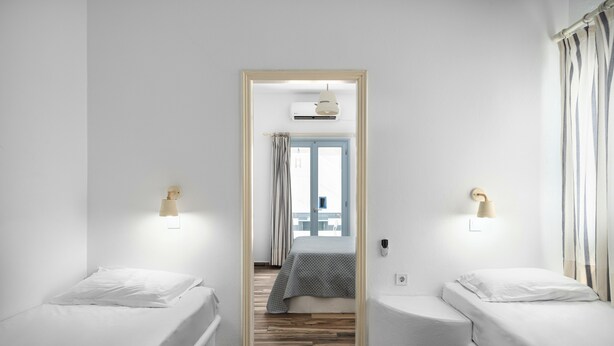Habitació d'hotel amb dos llits individuals, un terra de fusta, una finestra amb cortines i una porta que condueix a una altra habitació.