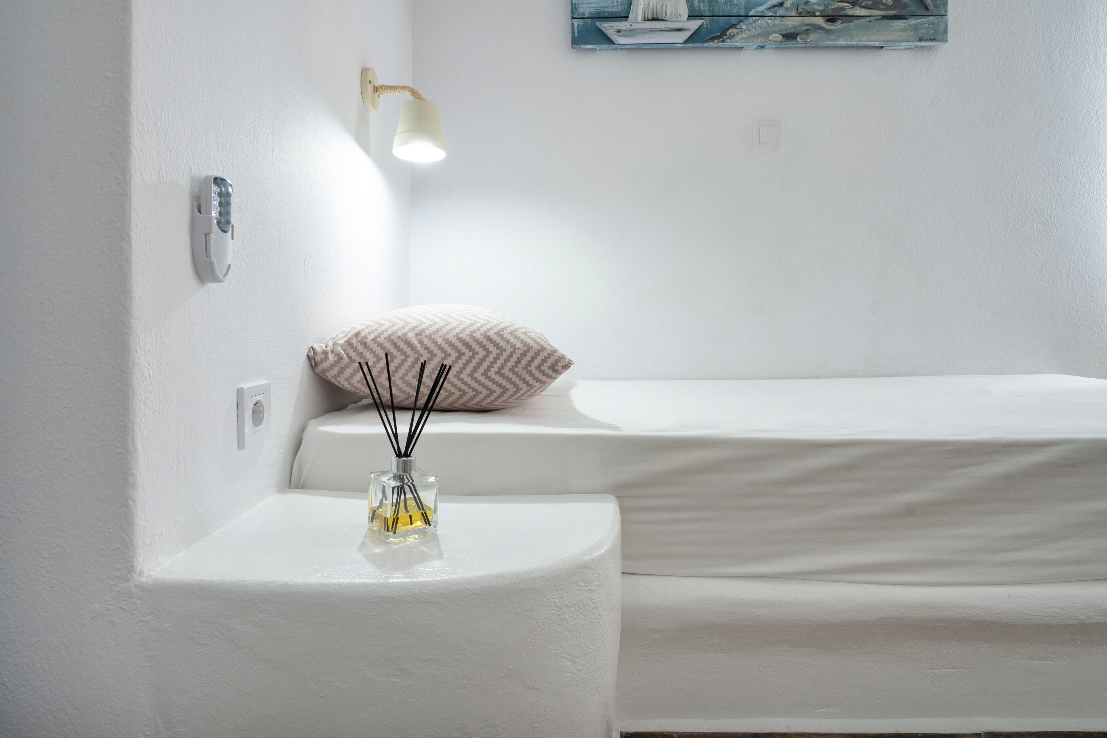 A white room with a bedside table, a lamp, a small vase with reeds, and a bed.