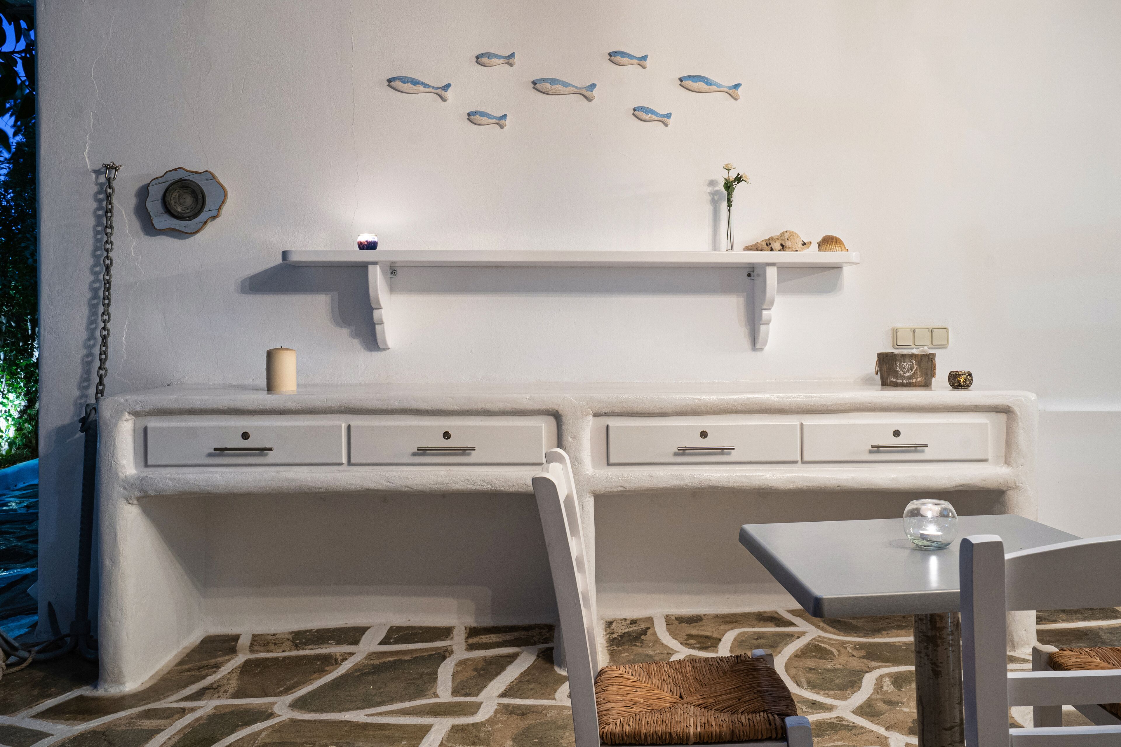Una habitación con un escritorio blanco, dos cajones, una silla blanca, una mesita pequeña y una estantería con adornos.