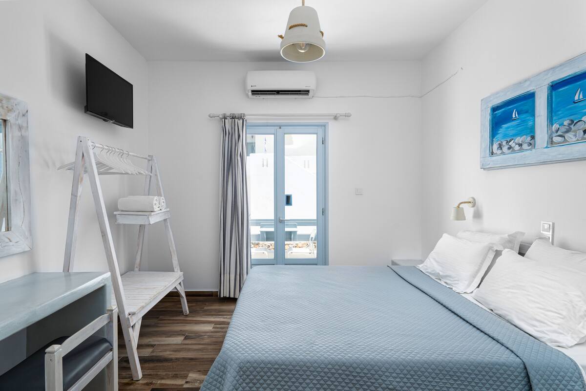 A bedroom with a bed, a TV, an air conditioner, a window with curtains, and a painting of sailboats.