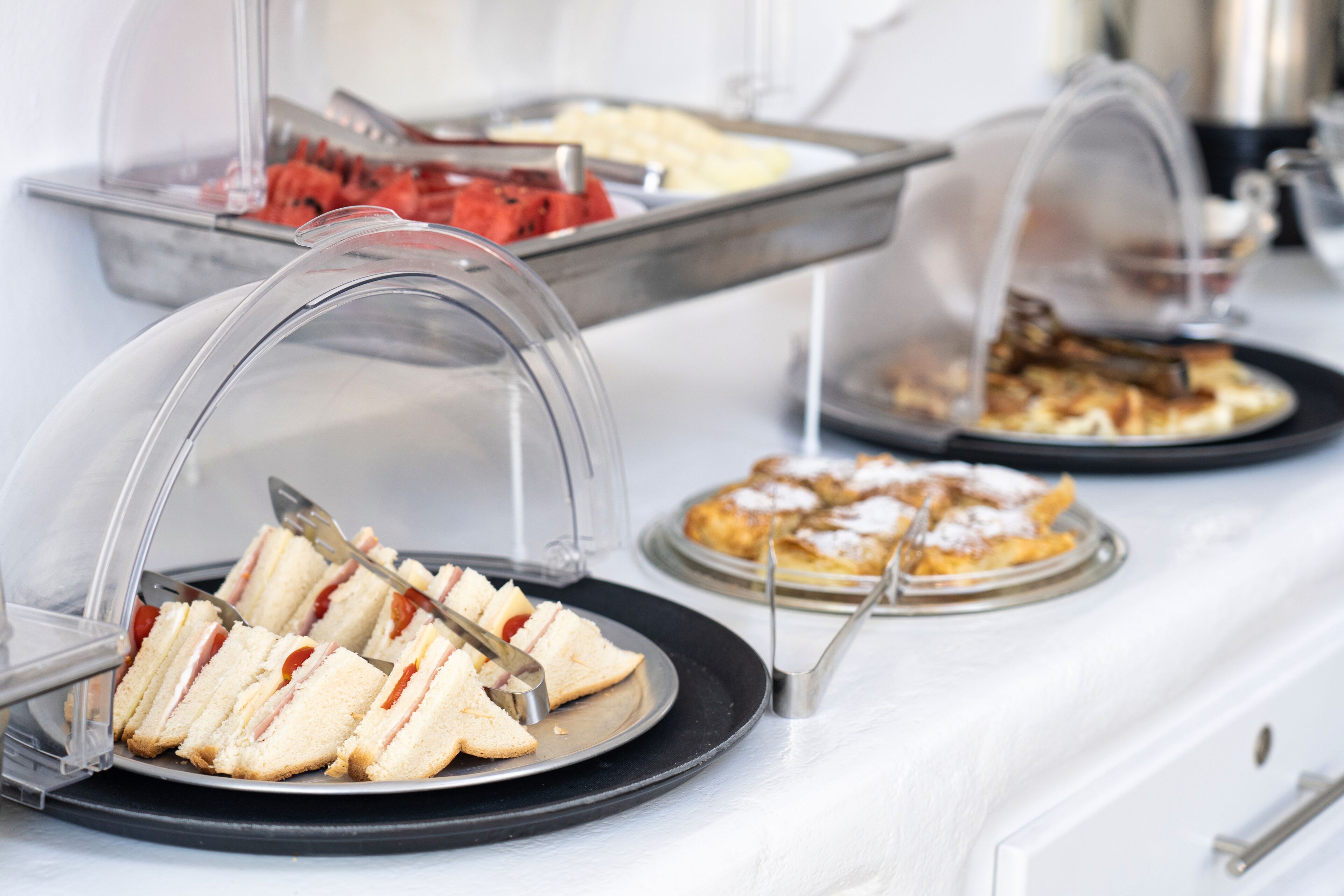 A buffet with various food items under clear covers, including sandwiches and pastries.
