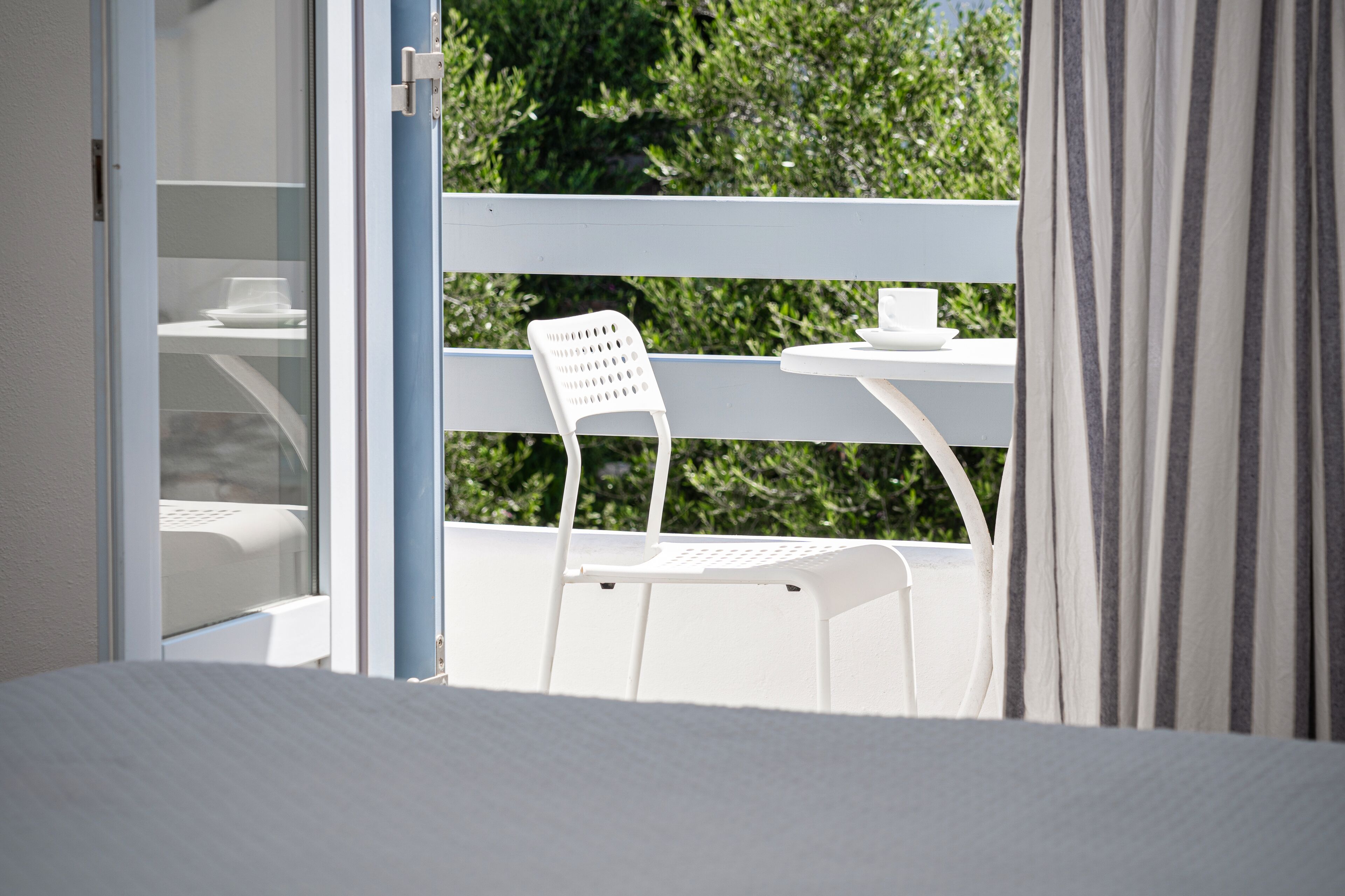 A white outdoor balcony with a table and chair, a glass door, and a view of greenery.