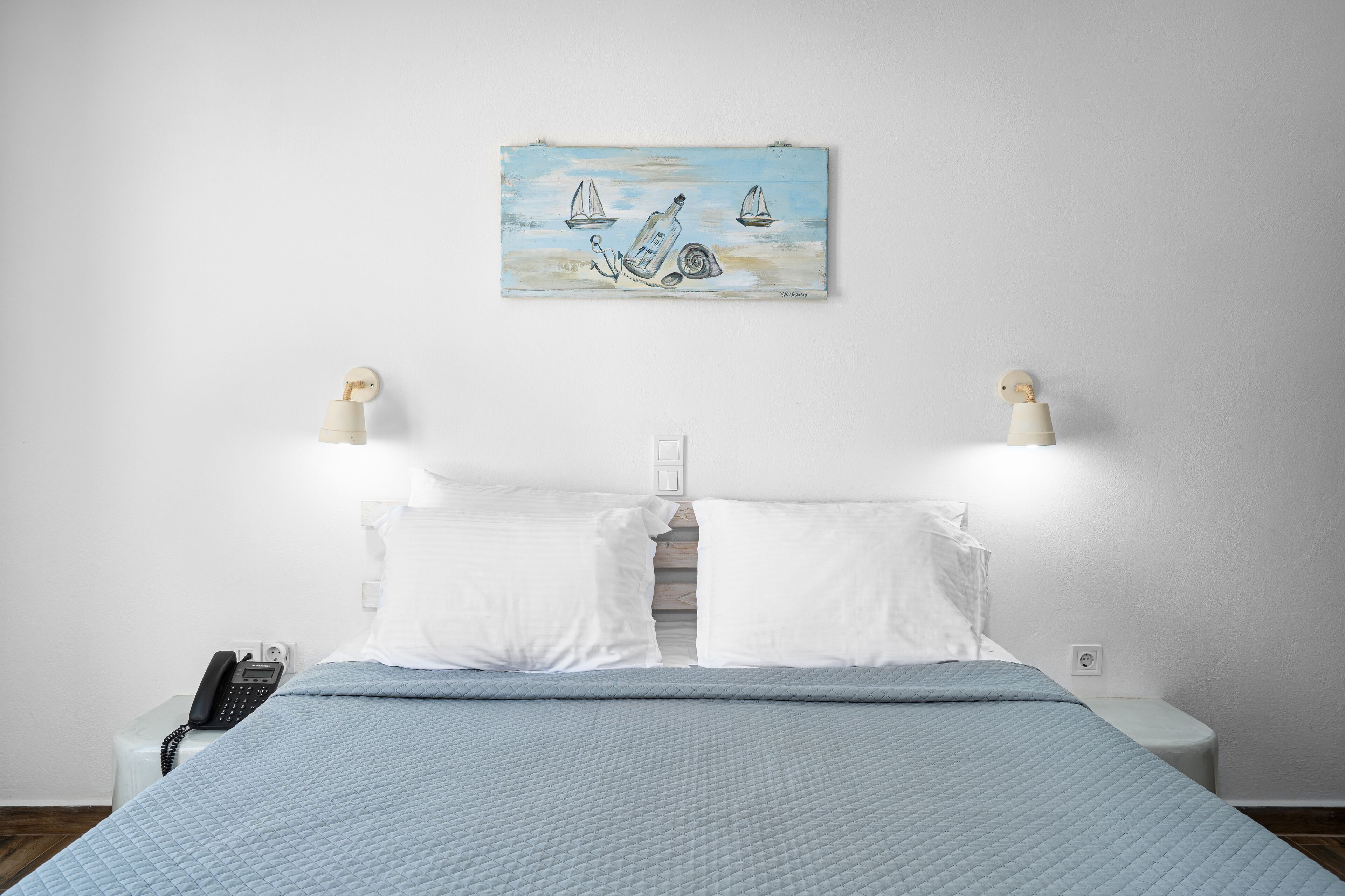 Ein ordentlich bezogenes Bett mit einer blauen Stegdecke, einem an der Wand hängenden Gemälde von Segelbooten und einem Nachttischtelefon.