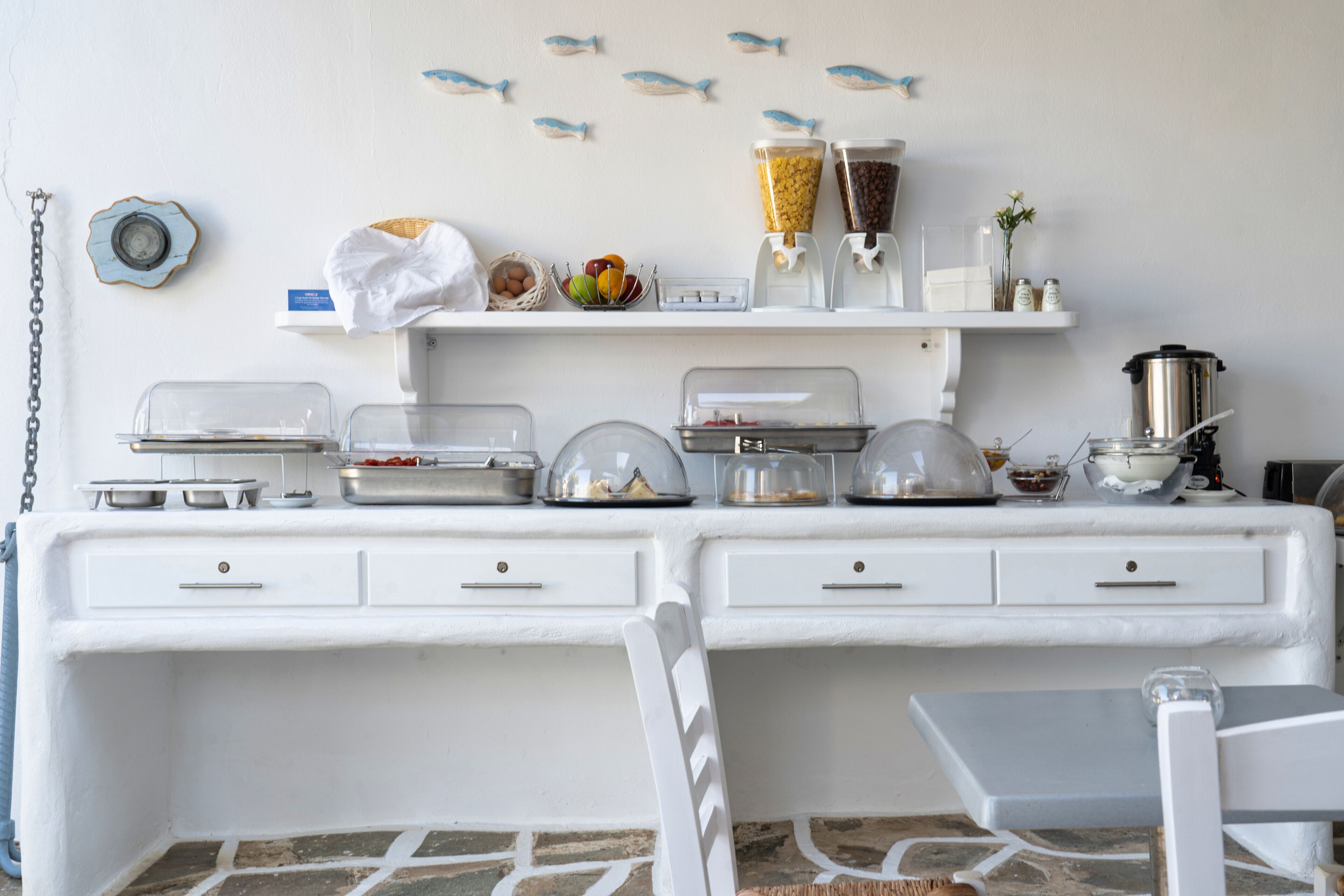 Disposição de buffet com diversos alimentos e recipientes sobre um balcão branco, uma cadeira e uma parede decorada com adesivos de peixes.