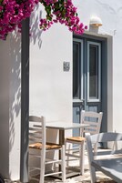 A white outdoor seating area with a table and chairs, a door with a number, and pink flowers hanging above.
