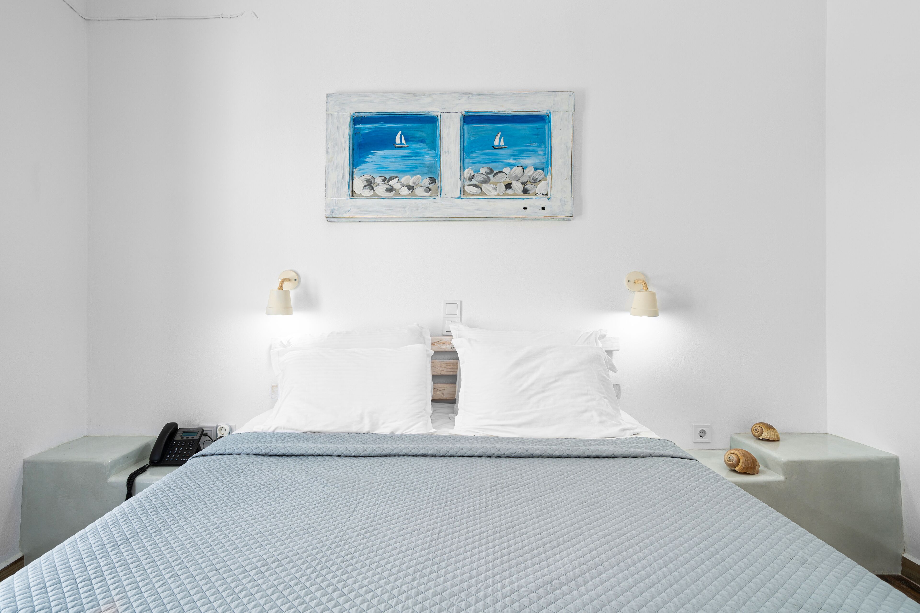 A bed with a quilted blue blanket, two white pillows, and a wall-mounted painting of sailboats.