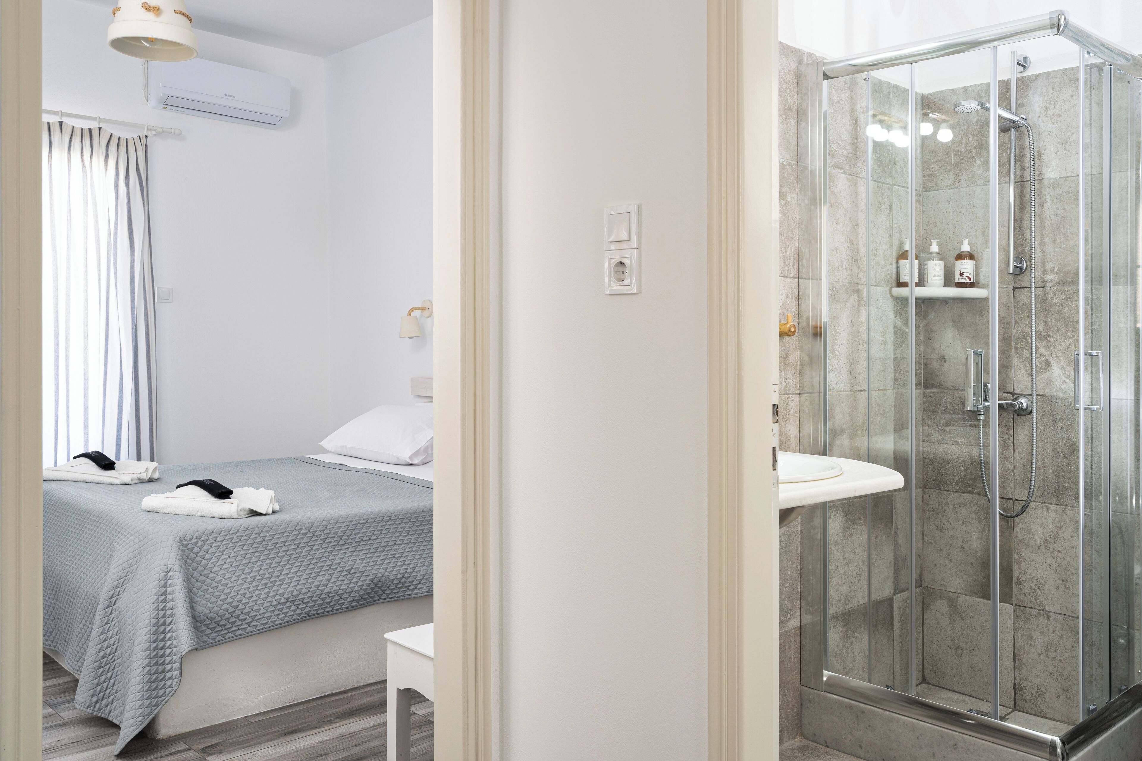 Quarto de hotel com cama, mesa de cabeceira, chuveiro com box de vidro e pia no banheiro.