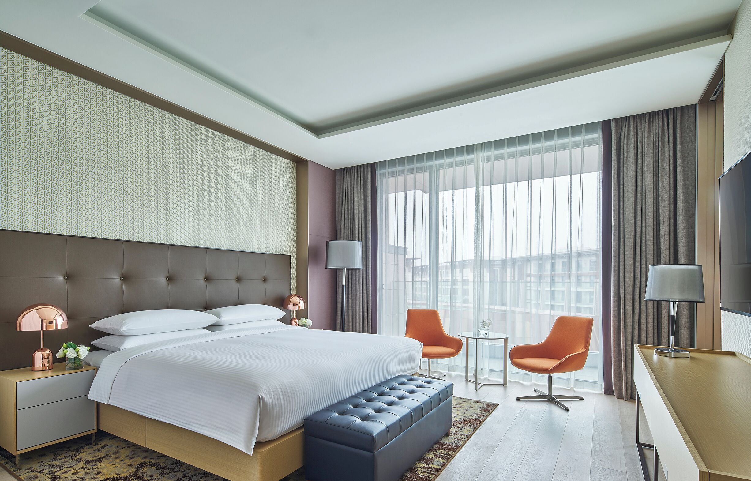 A modern hotel room with a large bed, a small desk, two chairs, and a large window with curtains.