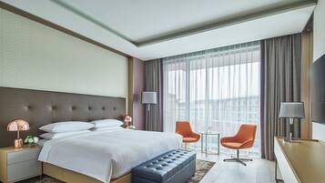 A modern hotel room with a large bed, a small desk, two chairs, and a large window with curtains.