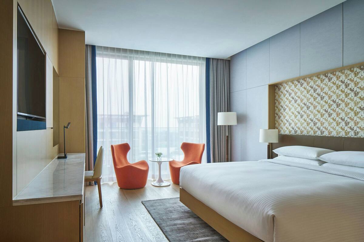 A hotel room with a large bed, a TV, two orange chairs, a small table, and a view of the city through large windows.