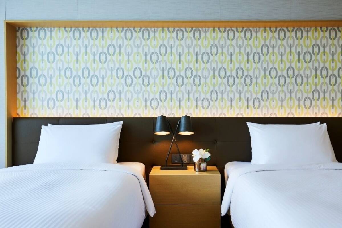 A hotel room with two beds, a wooden headboard, a patterned wallpaper, and a nightstand with a lamp and a vase with flowers.