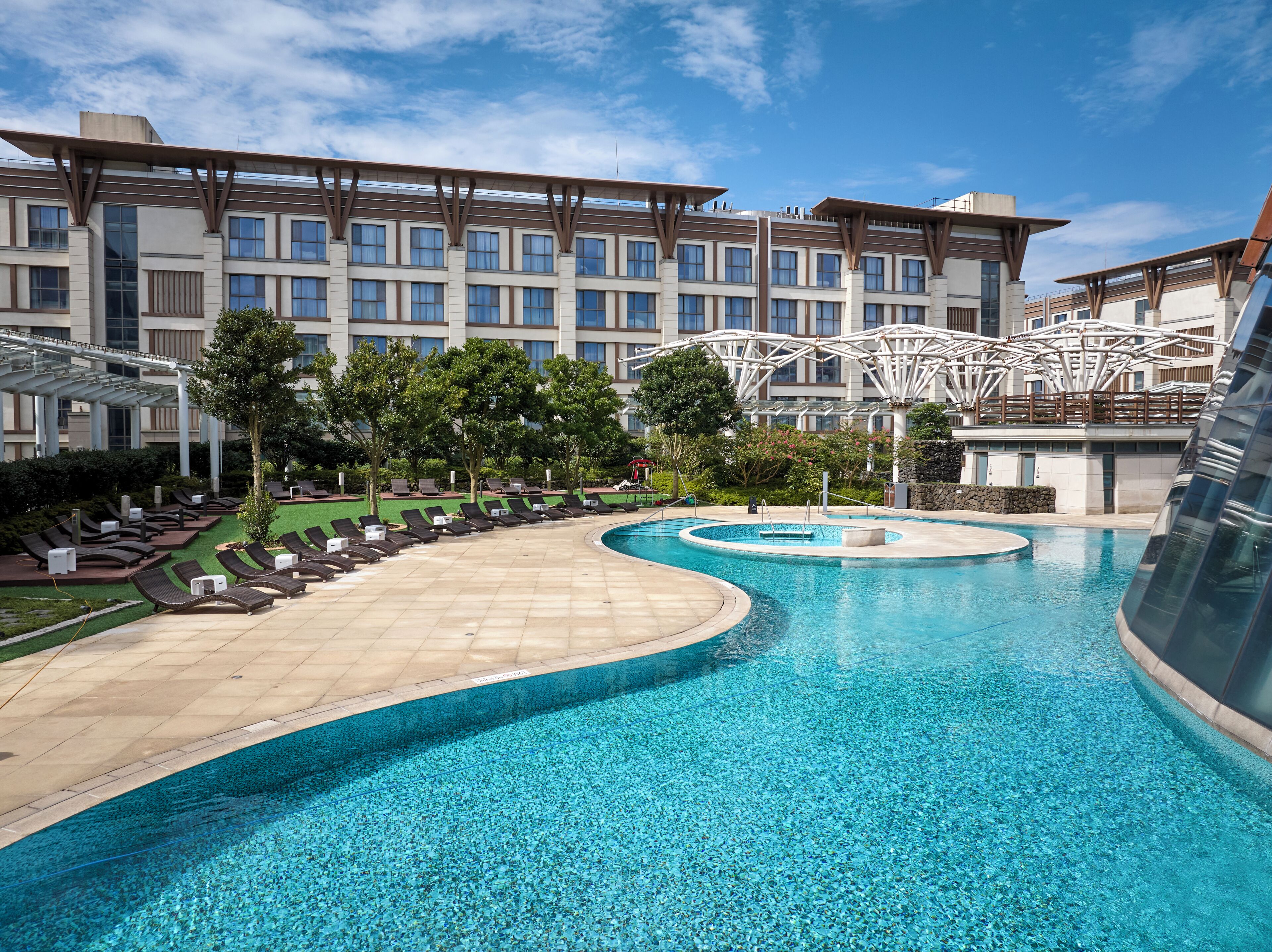 A hotel with a large pool, sun loungers, and a modern building with a unique roof structure.