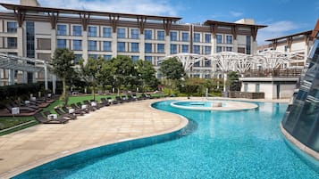A hotel with a large pool, sun loungers, and a modern building with a unique roof structure.