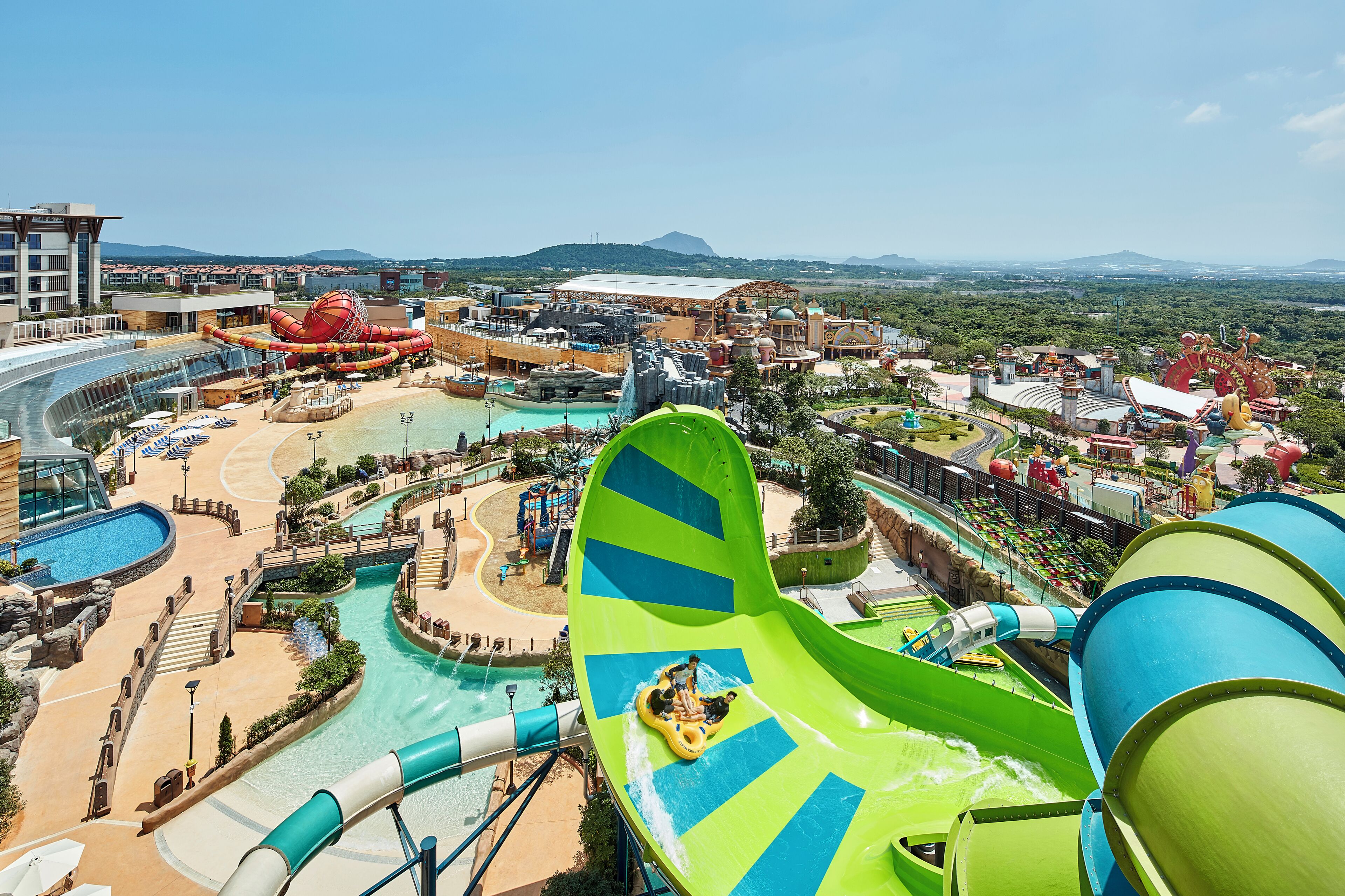 A water park with a large green water slide, a pool area, and a mountainous backdrop.