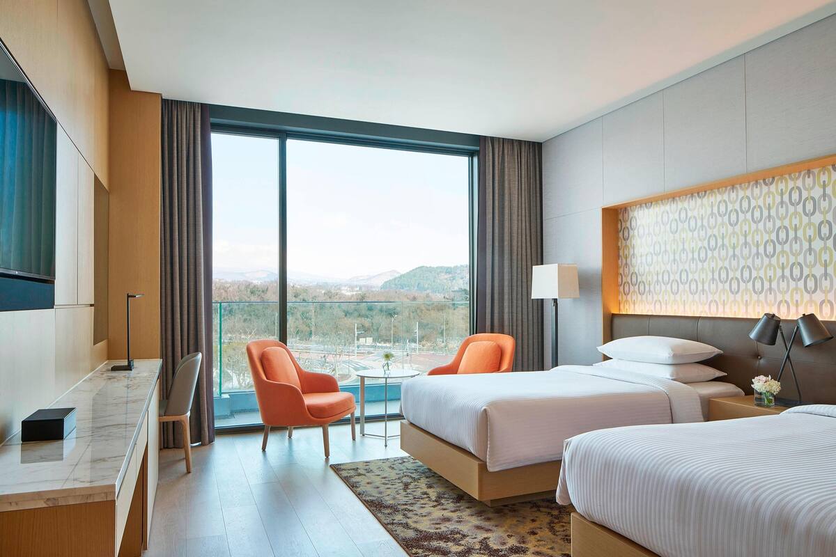 A hotel room with two beds, a desk, a chair, a small table, and a large window offering a view of the outdoors.