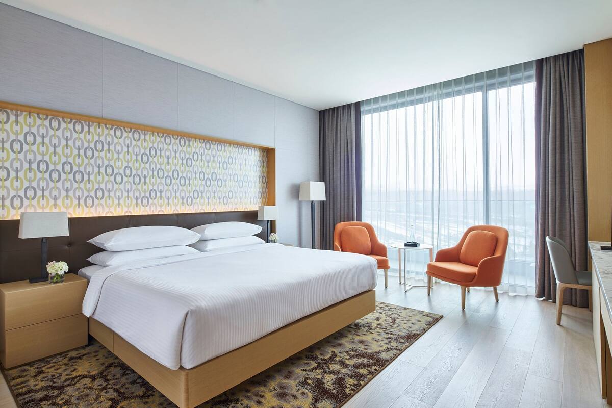 A modern hotel room with a large bed, two armchairs, a desk, and a view of the city through large windows.