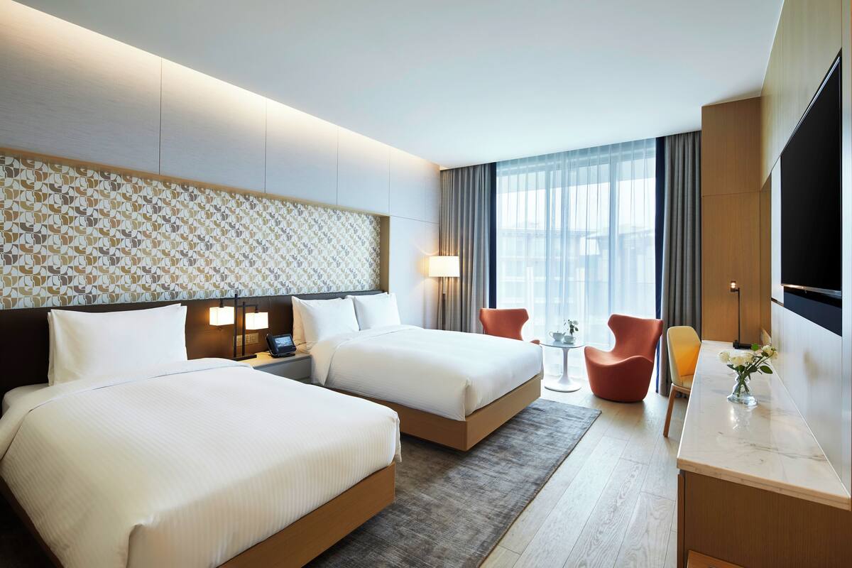 A hotel room with two beds, a desk, a TV, and a large window with curtains.