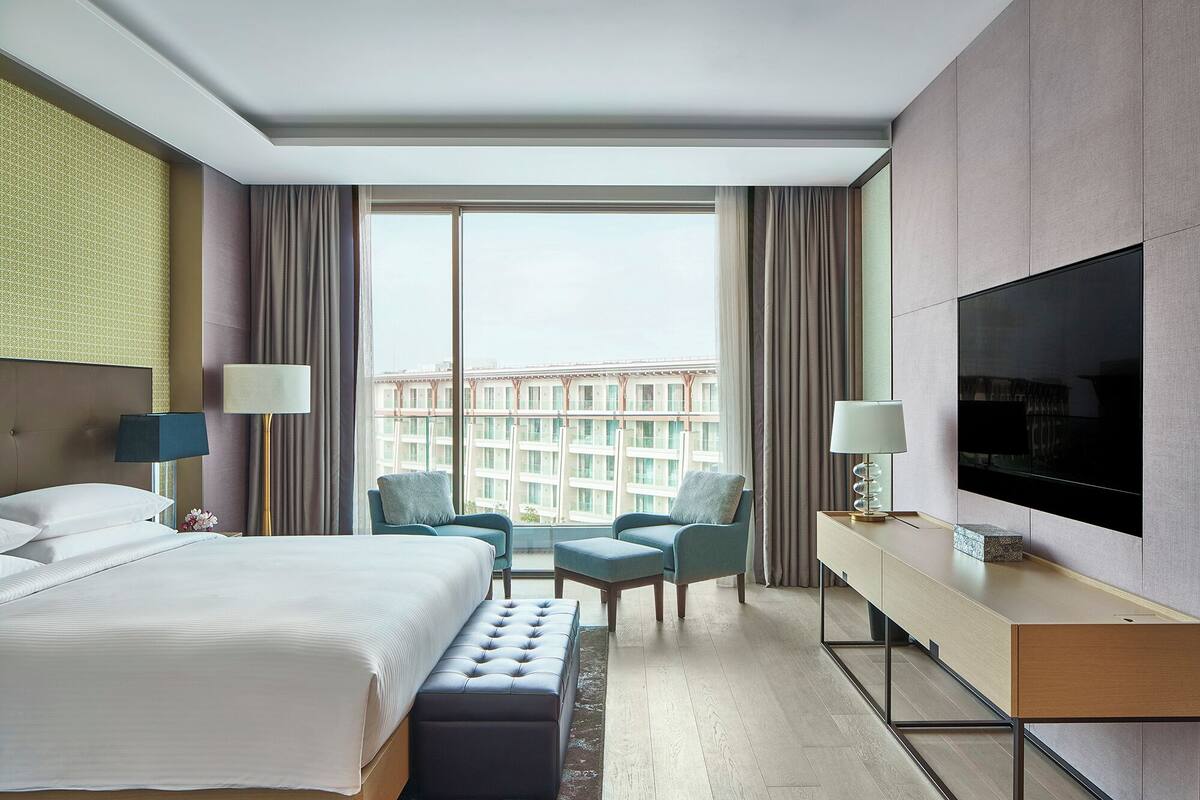 A hotel room with a bed, two armchairs, a TV, and a large window with a city view.