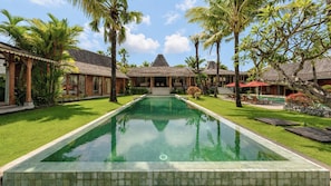 A pool with a tiled edge, surrounded by palm trees and a building with a thatched roof. - Villa Mannao by Villa Finder (Kerobokan)