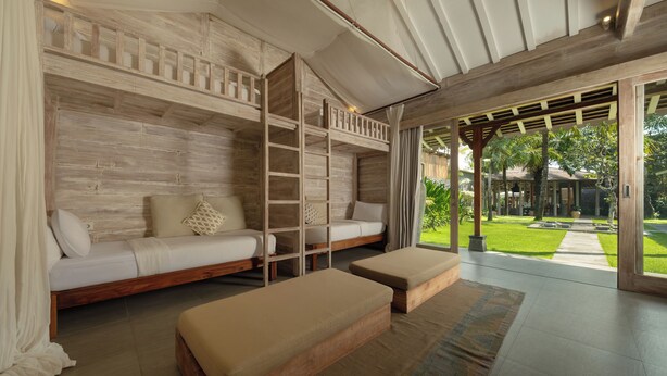 A room with a wooden wall, a sofa, a bed, and a view of a garden. - Villa Mannao by Villa Finder (Kerobokan)