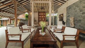 A covered outdoor seating area with wooden furniture, a central table with a vase of flowers, and a stone wall. - Villa Mannao by Villa Finder (Kerobokan)
