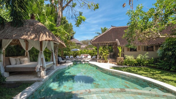 Uma área de piscina com um gazebo de telhado de palha, espreguiçadeiras e piscina cristalina.