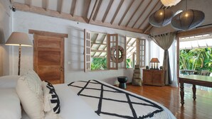 Quarto com cama, vigas de madeira no teto, porta de madeira, janela com cortinas, mesa de cabeceira e vista para o verde lá fora.