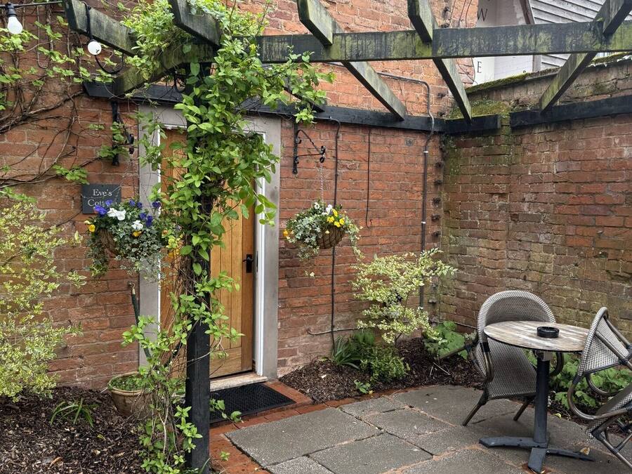 A brick building with a wooden door, a small garden with a table and chairs, and hanging flower baskets.