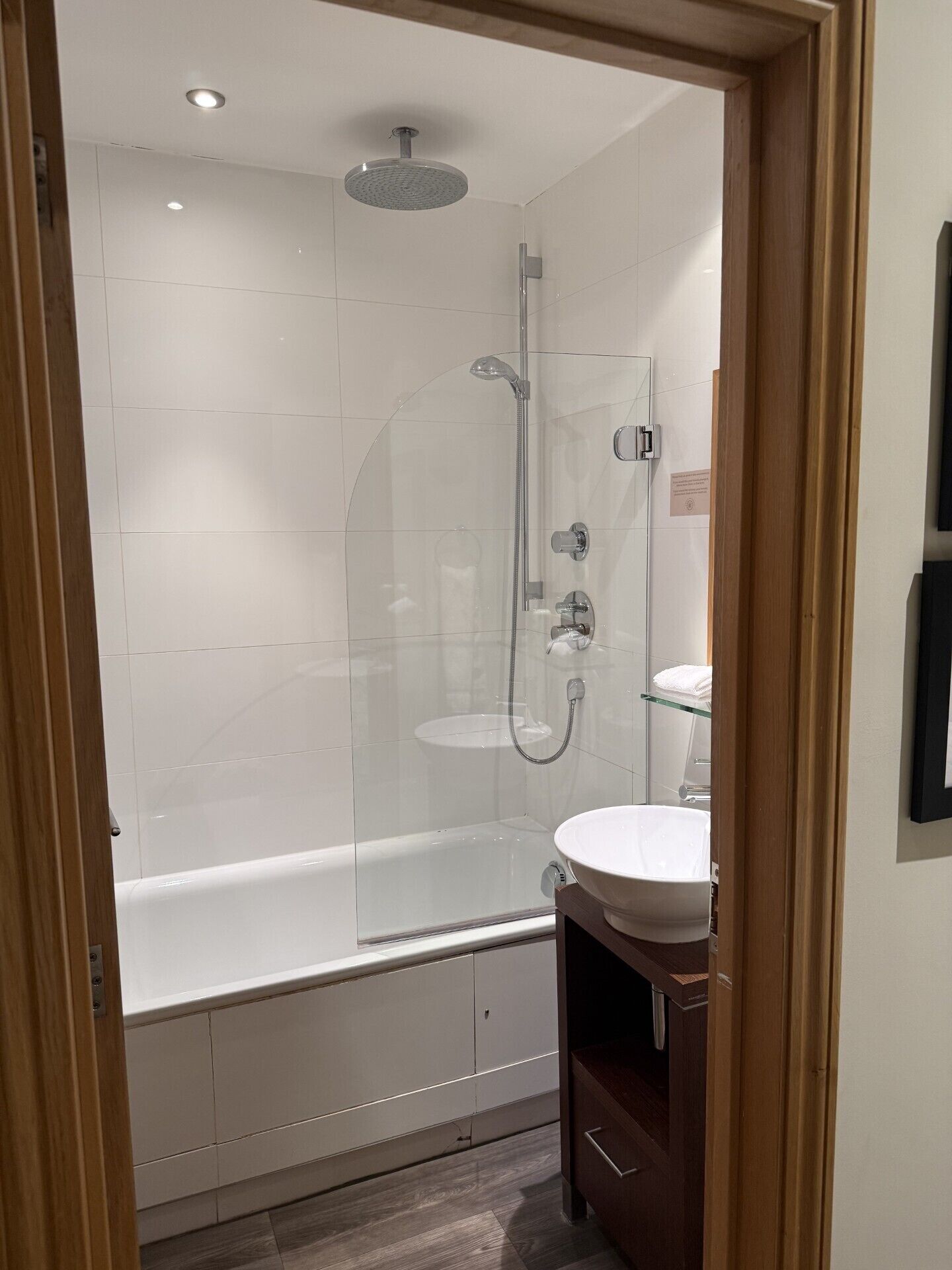 Un baño moderno con ducha de vidrio, bañera blanca y tocador de madera con lavamanos.
