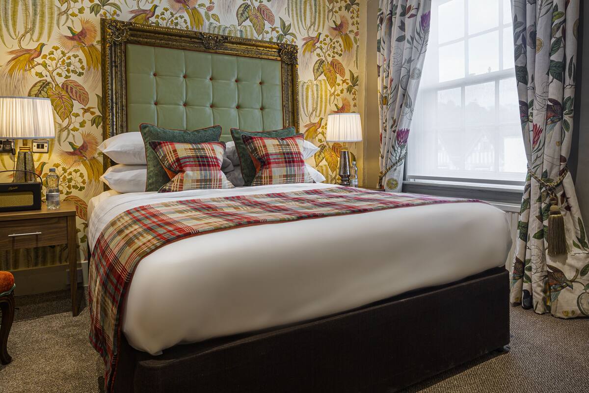 A well-appointed hotel room with a large bed, a tufted headboard, floral wallpaper, and a window with curtains.