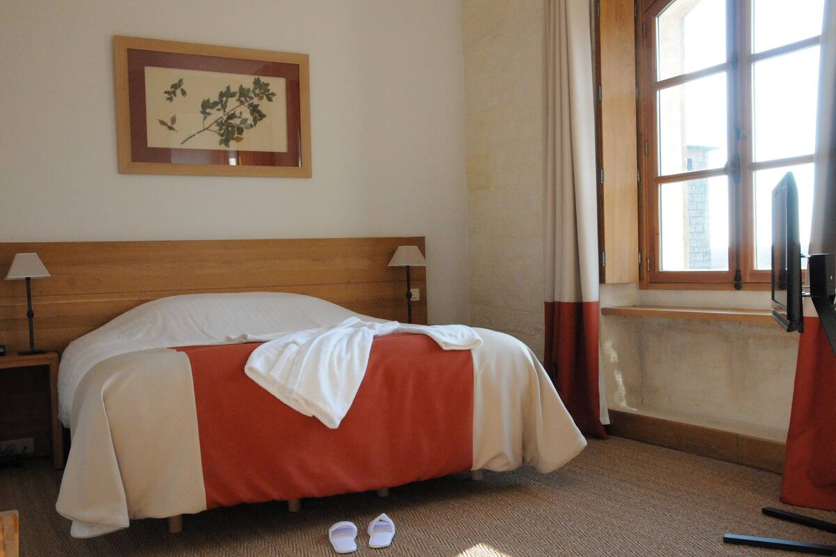 Une chambre d’hôtel avec une tête de lit en bois, un lit recouvert d’une couette rouge et blanche, une paire de pantoufles posées sur le sol, une estampe botanique encadrée accrochée au mur et une fenêtre avec des rideaux.