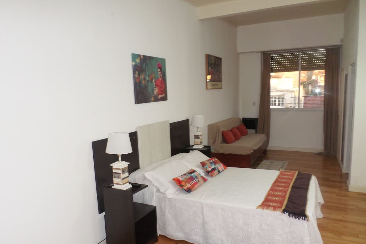A bedroom with a bed, bedside tables, a nightstand, a window with blinds, and a door leading to another room.