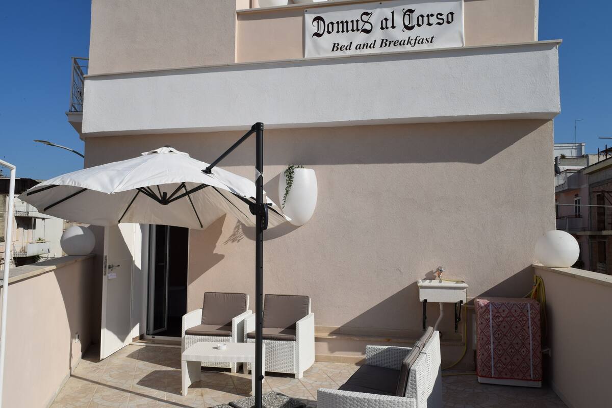A balcony with white wicker furniture, a white umbrella, and a sign reading "Domus al Corso Bed and Breakfast".