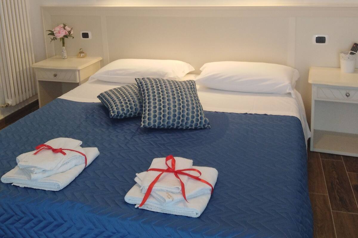 A neatly made bed with blue bedding, white pillows, and towels with red ribbons. There are wall-mounted pictures depicting landscapes and architecture.