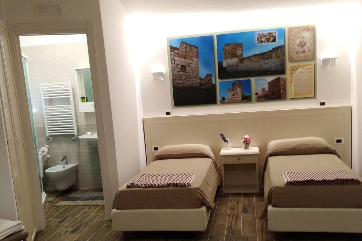 A hotel room with two beds, a bedside table with a lamp, a bathroom with a bidet, and a wooden floor.