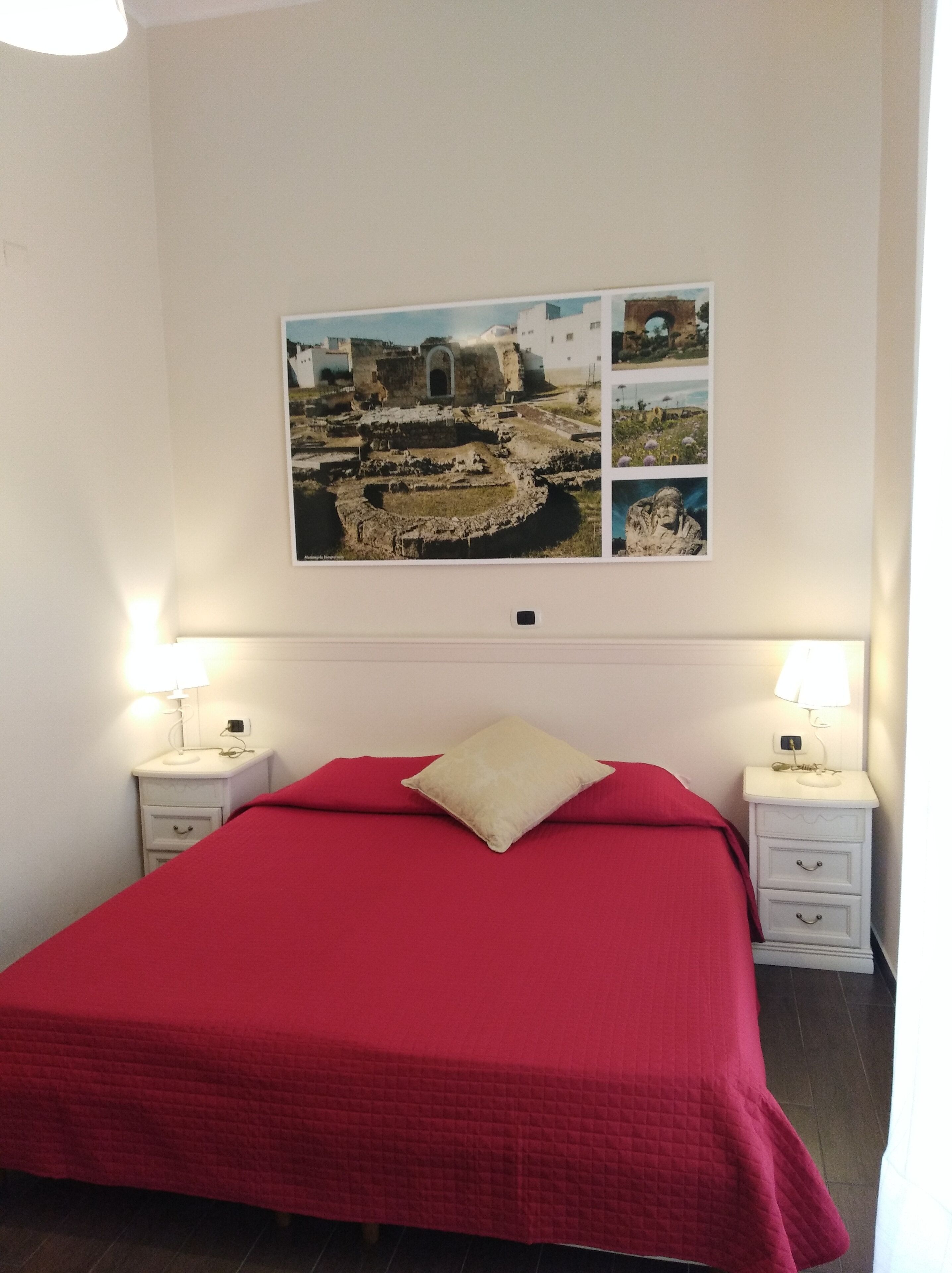 Quarto com colcha vermelha, duas mesas de cabeceira com abajures e um quadro na parede retratando um sítio arqueológico.
