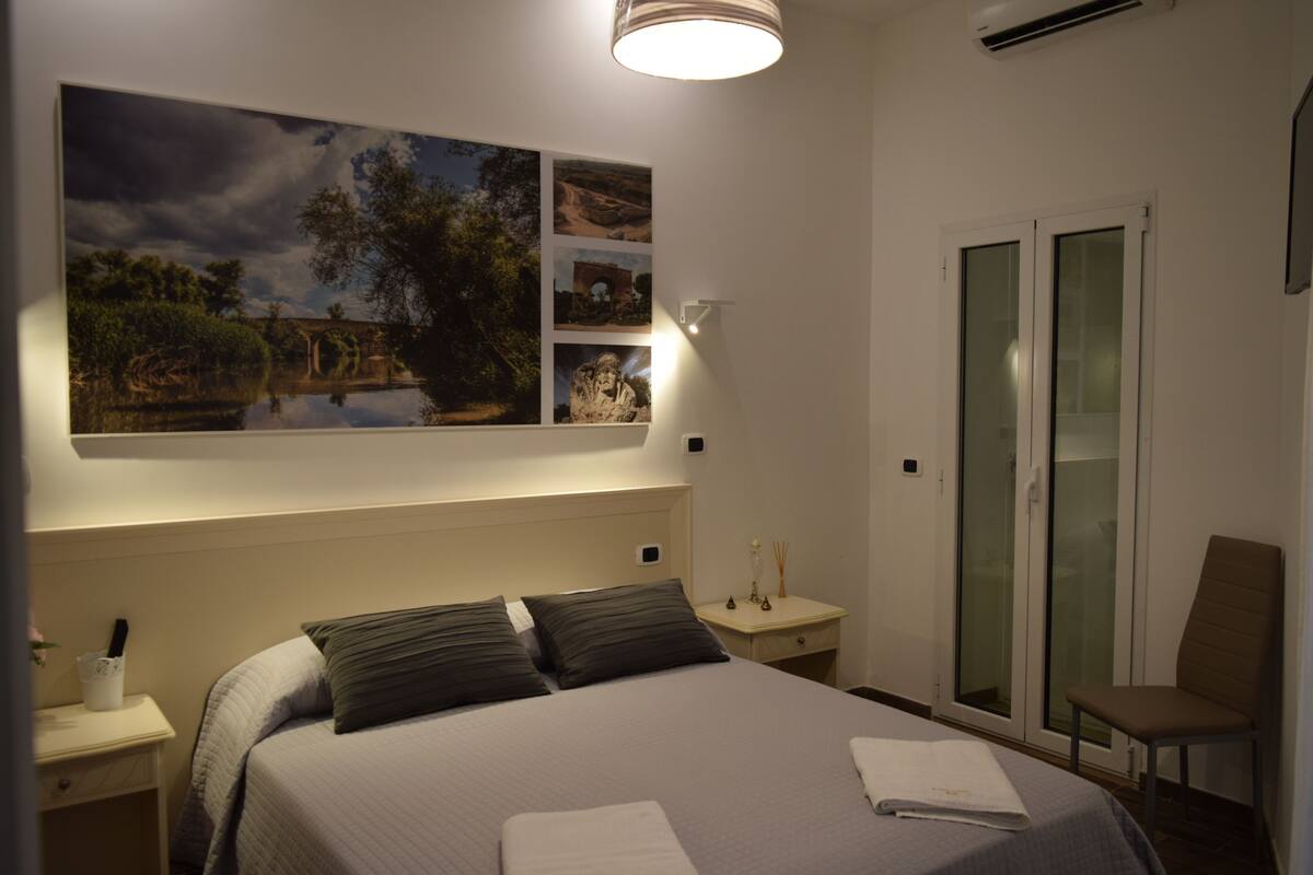 A bedroom with a large bed, a nightstand, a chair, a wall-mounted lamp, and a sliding glass door.